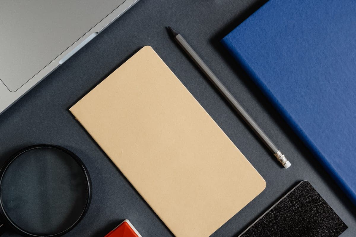 A tan notebook, a pencil, a blue notebook and a round magnifier laid out on a dark gray desk